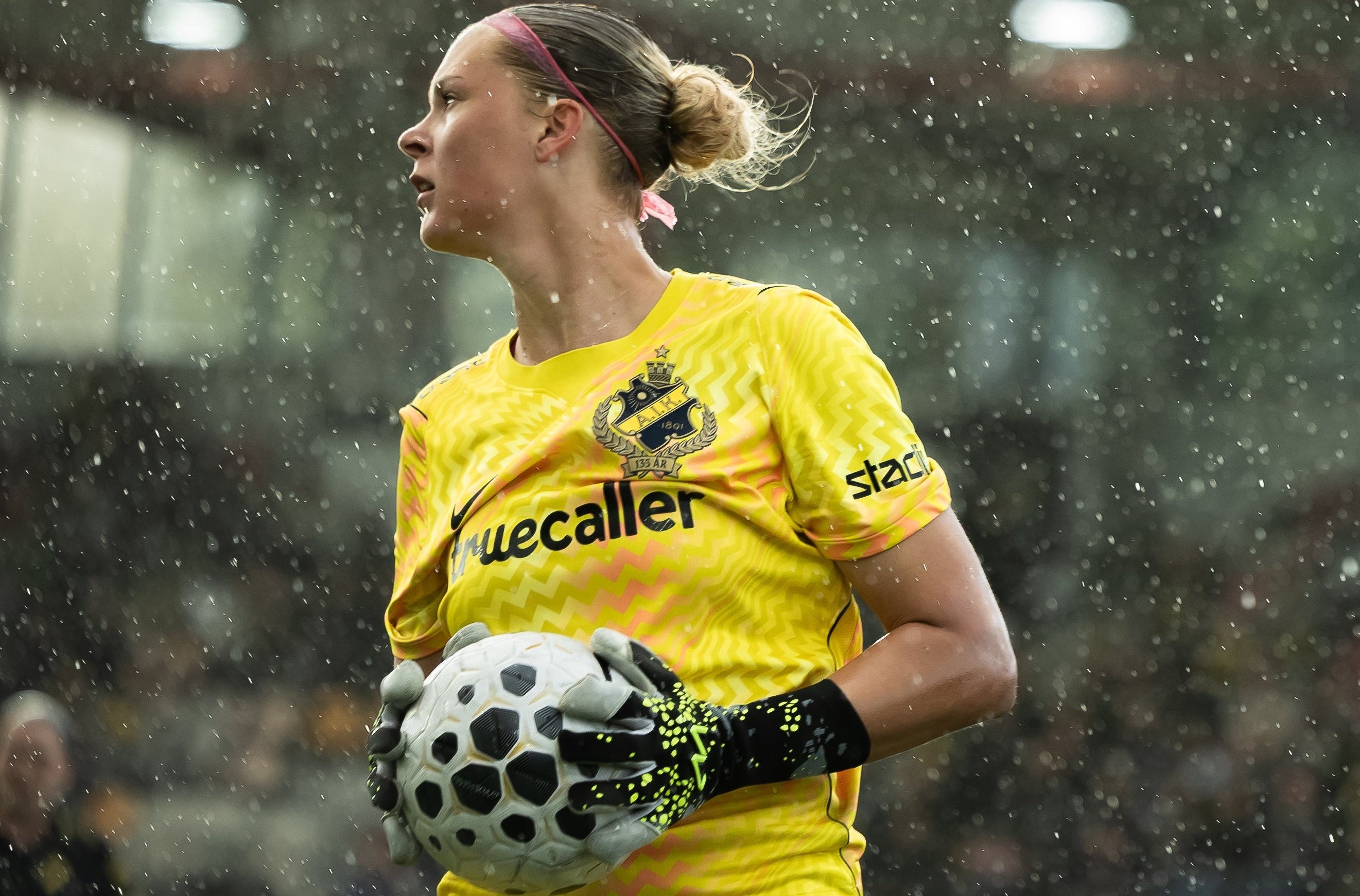 A female soccer goalkeeper in a bright yellow jersey with 'uecaller' and 'staeck' logos, holding a soccer ball in the rain. She has a pink headband and AM gloves Revontulet black-and-green goalkeeper gloves.